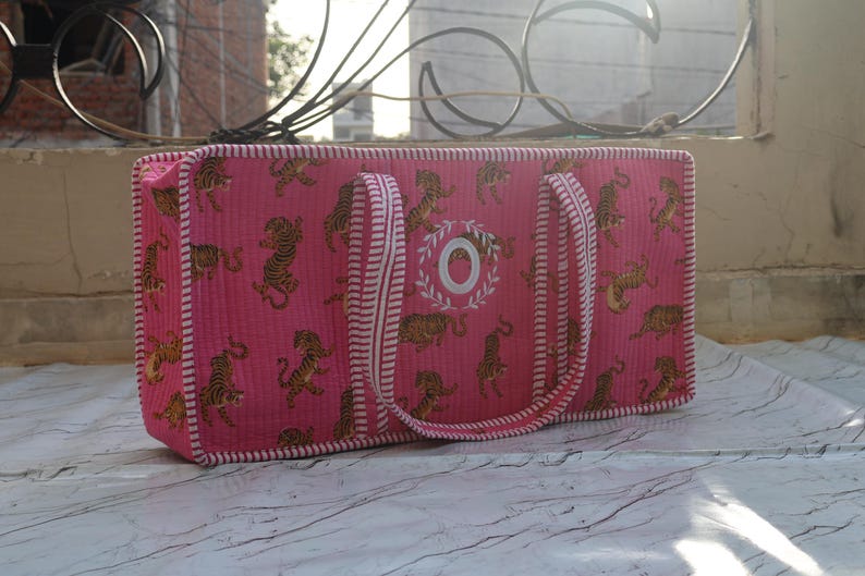 Puede incluir: Un bolso rectangular acolchado de color rosa con un patr&oacute;n repetitivo de tigres en tonos marrones y negros. El bolso tiene bordes y asas a rayas blancas y rosas, con una letra O bordada en blanco rodeada por un dise&ntilde;o de corona de laurel.