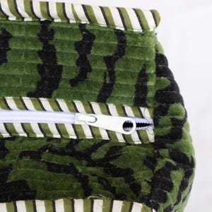 May include: Close-up of a green and black patterned fabric with a white zipper. The fabric has a textured, quilted appearance. The top and bottom edges are trimmed with a striped fabric in white and green.