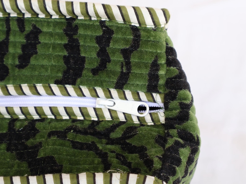 May include: Close-up of a green and black patterned fabric with a white zipper. The fabric has a textured, quilted appearance. The top and bottom edges are trimmed with a striped fabric in white and green.