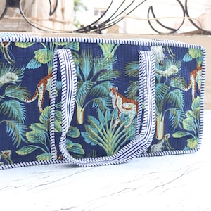 Puede incluir: Un bolso rectangular acolchado con fondo azul oscuro y un patr&oacute;n repetitivo de guepardos y hojas de palmera tropical en tonos verdes y azul verdoso. El bolso tiene dos asas a juego y bordes ribeteados con una tela de rayas blancas y azules.