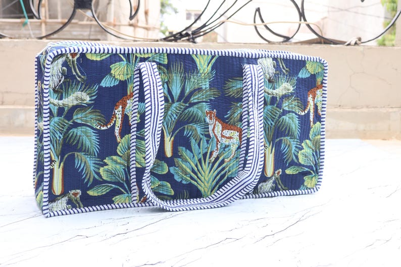 Puede incluir: Un bolso rectangular acolchado con fondo azul oscuro y un patr&oacute;n repetitivo de guepardos y hojas de palmera tropical en tonos verdes y azul verdoso. El bolso tiene dos asas a juego y bordes ribeteados con una tela de rayas blancas y azules.