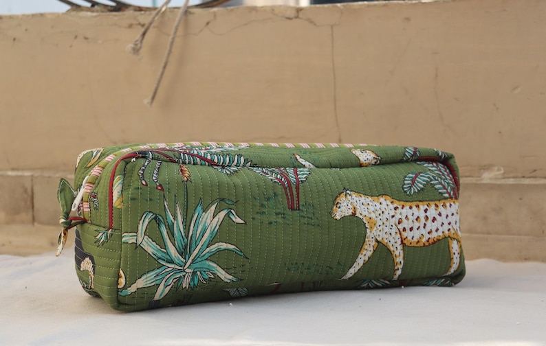 May include: Green rectangular cosmetic bag with a quilted texture. The bag features a jungle-themed print with white and yellow leopards, and green and blue plants. It has a zipper closure and a small fabric loop on the side.