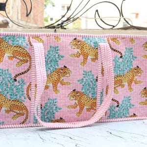 Puede incluir: Un bolso tote rectangular acolchado con un patr&oacute;n repetitivo de leopardos amarillos y negros intercalados con follaje verde azulado sobre un fondo rosa claro. El bolso tiene dos asas y un borde de tela a rayas rosas y blancas.