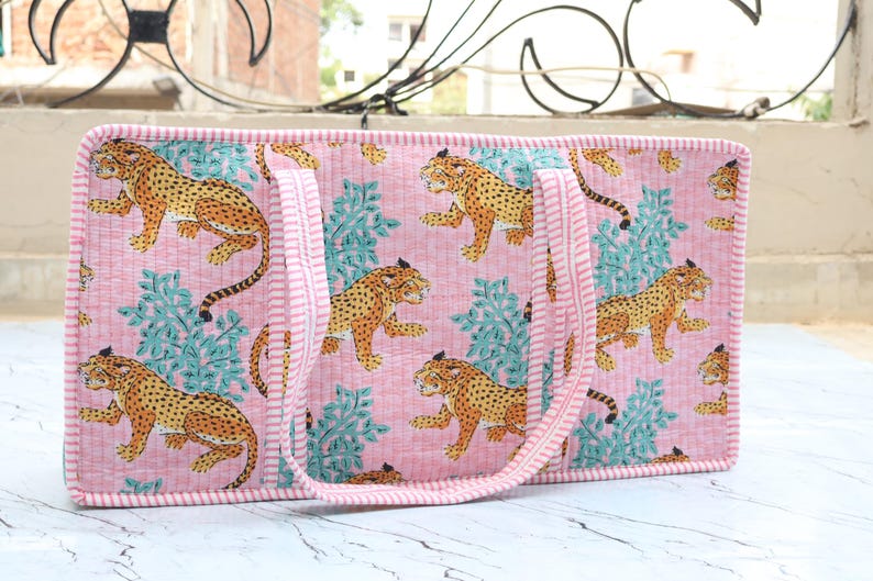 Puede incluir: Un bolso tote rectangular acolchado con un patr&oacute;n repetitivo de leopardos amarillos y negros intercalados con follaje verde azulado sobre un fondo rosa claro. El bolso tiene dos asas y un borde de tela a rayas rosas y blancas.