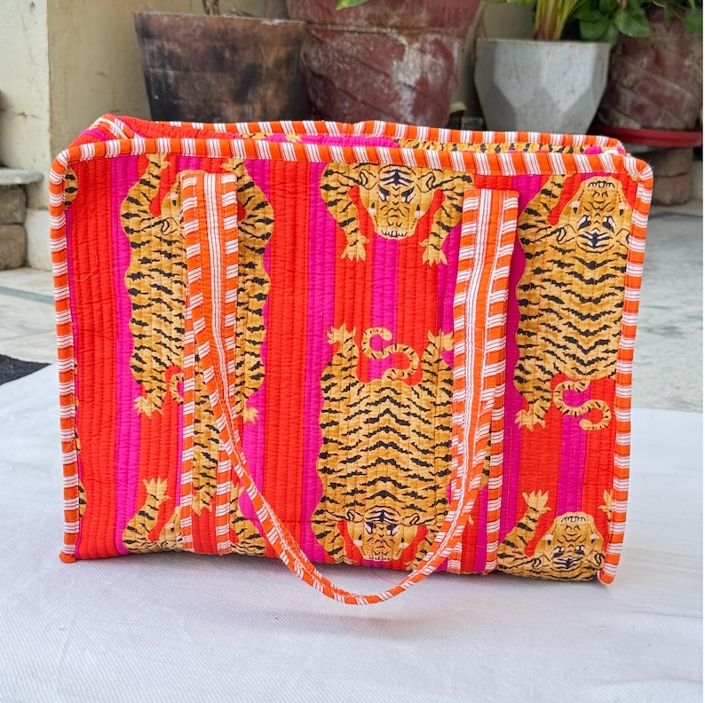 May include: A rectangular tote bag with a vibrant design. The bag features a repeating pattern of orange and black tiger illustrations set against a background of vertical stripes in pink and orange. The bag has a matching orange handle.