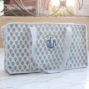 Puede incluir: Un bolso tote acolchado rectangular con un patr&oacute;n floral repetido en azul y verde sobre un fondo blanco. El bolso tiene bordes y asas de cuadros vichy en azul claro y blanco, con un monograma circular bordado que dice ALN en color azul.