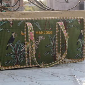 Puede incluir: Un bolso rectangular de tela acolchada en color verde oliva con un estampado de selva tropical que incluye tucanes, cebras y palmeras. La palabra MAHJONG est&aacute; bordada en amarillo en la parte frontal. El bolso tiene dos asas de tela a rayas.