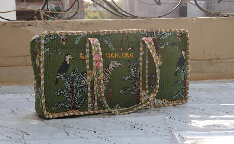 Puede incluir: Un bolso rectangular de tela acolchada en color verde oliva con un estampado de selva tropical que incluye tucanes, cebras y palmeras. La palabra MAHJONG est&aacute; bordada en amarillo en la parte frontal. El bolso tiene dos asas de tela a rayas.