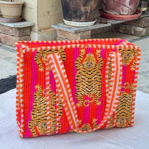 May include: A rectangular tote bag with a striking design. The bag showcases a repeating pattern of orange and black tiger illustrations set against vertical pink and orange stripes. It has orange and white striped handles.