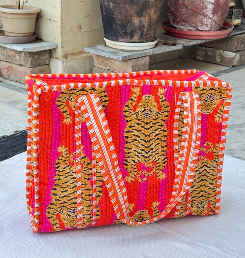May include: A rectangular tote bag with a striking design. The bag showcases a repeating pattern of orange and black tiger illustrations set against vertical pink and orange stripes. It has orange and white striped handles.