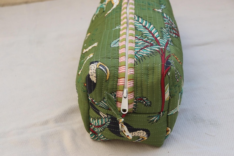 May include: Green quilted fabric bag with a tropical print featuring toucans, trees, and other wildlife. The bag has a white zipper with pink and white striped trim. The bag is rectangular and appears to be a cosmetic or travel bag.