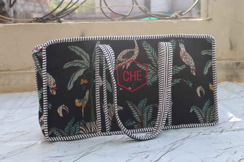 Puede incluir: Un bolso rectangular negro con un estampado tropical de palmeras, p&aacute;jaros y cebras. El bolso tiene asas y bordes a rayas blancas y negras. En la parte delantera hay un hex&aacute;gono rojo bordado que contiene las letras CHE en su interior.