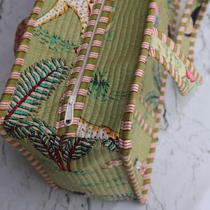 Puede incluir: Un bolso de tela acolchada en tono verde oliva claro con un patr&oacute;n repetitivo de hojas de palmera tropicales y animales. El bolso incluye un cierre de cremallera blanco en la parte superior y est&aacute; rematado con ribetes a rayas en rosa y blanco.