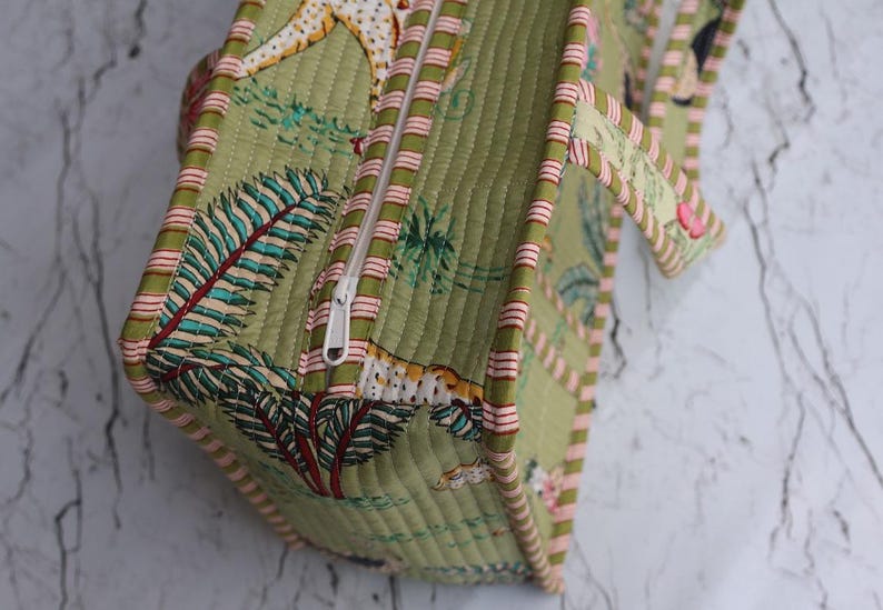 Puede incluir: Un bolso de tela acolchada en tono verde oliva claro con un patr&oacute;n repetitivo de hojas de palmera tropicales y animales. El bolso incluye un cierre de cremallera blanco en la parte superior y est&aacute; rematado con ribetes a rayas en rosa y blanco.