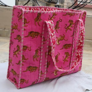 May include: A quilted tote bag in a vibrant pink hue, decorated with a repeating pattern of orange and black tiger illustrations. The bag has sturdy handles and a contrasting patterned interior, ideal for carrying essentials.