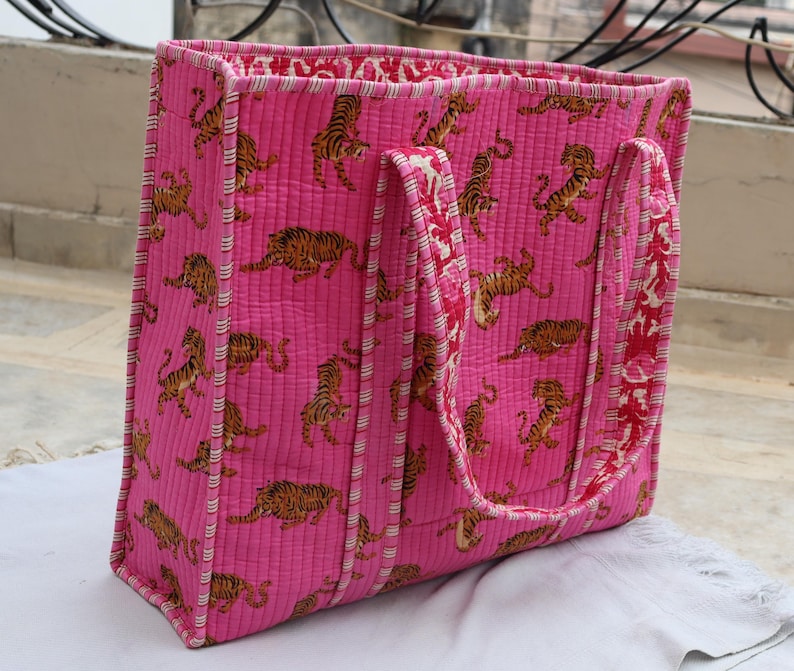 May include: A quilted tote bag in a vibrant pink hue, decorated with a repeating pattern of orange and black tiger illustrations. The bag has sturdy handles and a contrasting patterned interior, ideal for carrying essentials.