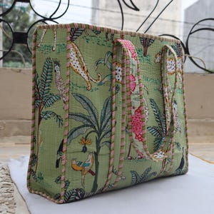 May include: A large, rectangular tote bag with a green jungle print featuring leopards, birds, and foliage. The bag has pink and white striped trim and matching handles. The bag is made of a quilted fabric.