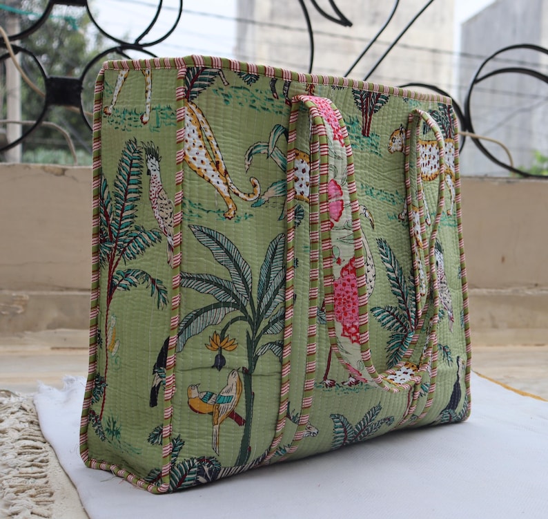 May include: A large, rectangular tote bag with a green jungle print featuring leopards, birds, and foliage. The bag has pink and white striped trim and matching handles. The bag is made of a quilted fabric.