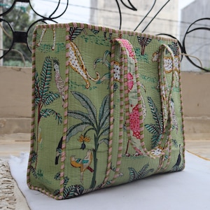 May include: Green tote bag with a jungle print featuring leopards, birds, and tropical plants. The bag has a rectangular shape with two handles and is trimmed with pink and green striped fabric. The bag is made of quilted fabric.