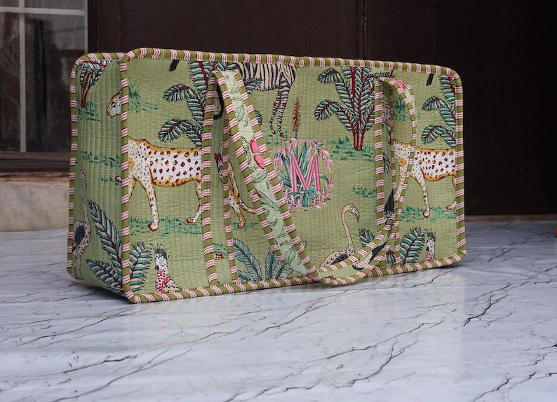 Puede incluir: Un bolso tote acolchado rectangular de tela verde claro con un estampado de selva que incluye guepardos, cebras y aves. El bolso tiene dos asas de tela a juego y un borde a rayas rojas y blancas. Una letra M de color rosa est&aacute; bordada en el centro.