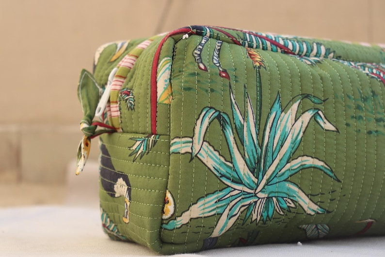 May include: Green quilted cosmetic bag with a botanical print featuring agave plants and other tropical elements. The bag has a rectangular shape with a zipper closure and a small fabric loop on the side.