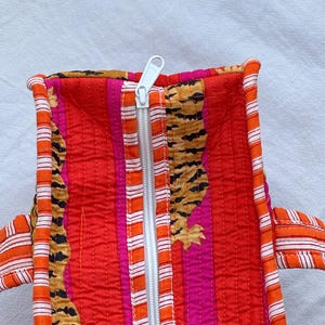 May include: Quilted tote bag with a vibrant tiger stripe and floral pattern in red, orange, and pink. The bag features a white zip and orange and white striped handles. The bag is made of a quilted fabric.