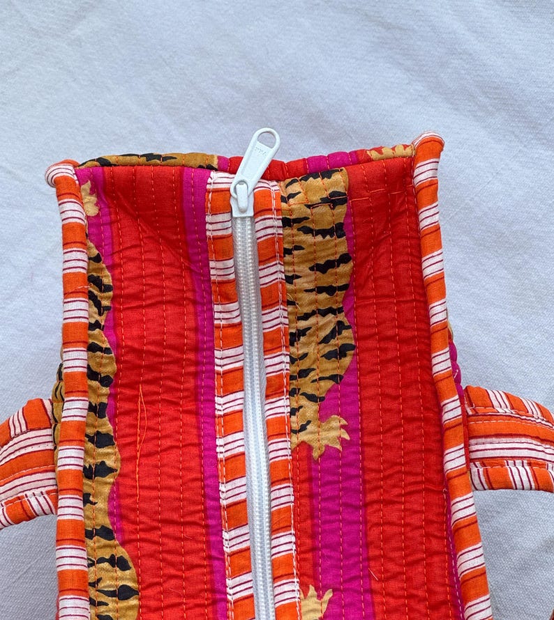 May include: Quilted tote bag with a vibrant tiger stripe and floral pattern in red, orange, and pink. The bag features a white zip and orange and white striped handles. The bag is made of a quilted fabric.