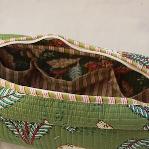 May include: Open green quilted bag with a white zipper and a floral pattern. The interior features multiple pockets and a striped fabric lining. The bag's exterior has a repeating pattern of leaves and other botanical elements.