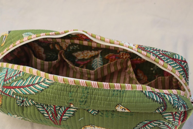 May include: Open green quilted bag with a white zipper and a floral pattern. The interior features multiple pockets and a striped fabric lining. The bag's exterior has a repeating pattern of leaves and other botanical elements.