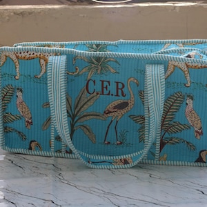 Puede incluir: Un bolso tote acolchado rectangular en color turquesa brillante con un patr&oacute;n repetitivo de aves tropicales, palmeras y guepardos. El bolso tiene dos asas a rayas y las letras C.E.R. bordadas en marr&oacute;n en el panel central delantero.
