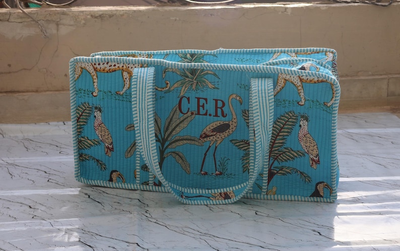 Puede incluir: Un bolso tote acolchado rectangular en color turquesa brillante con un patr&oacute;n repetitivo de aves tropicales, palmeras y guepardos. El bolso tiene dos asas a rayas y las letras C.E.R. bordadas en marr&oacute;n en el panel central delantero.