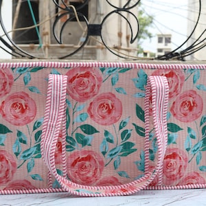 Puede incluir: Un bolso tote rectangular con fondo rosa claro y un patr&oacute;n repetitivo de rosas rojas y hojas verde azulado. El bolso tiene dos asas y los bordes est&aacute;n ribeteados con una tela a rayas rojas y blancas, lo que le da un aspecto estructurado.