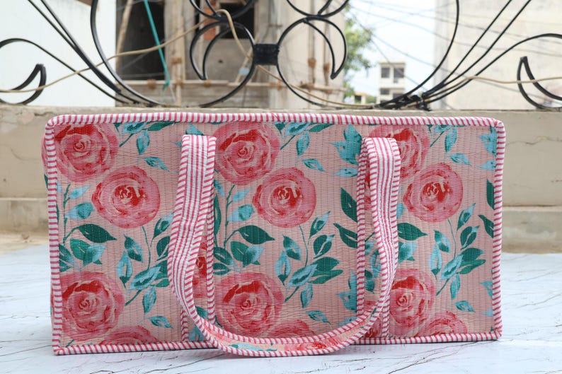 Puede incluir: Un bolso tote rectangular con fondo rosa claro y un patr&oacute;n repetitivo de rosas rojas y hojas verde azulado. El bolso tiene dos asas y los bordes est&aacute;n ribeteados con una tela a rayas rojas y blancas, lo que le da un aspecto estructurado.