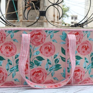 May include: A rectangular tote bag featuring a light pink background with a repeating pattern of red roses and teal green leaves. The bag has two handles and edges trimmed with a red and white striped fabric, creating a structured appearance.