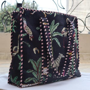 May include: Black tote bag with a quilted design featuring a jungle scene with parrots, flamingos, zebras, and palm trees. The bag has two handles and is trimmed with a pink and white striped border.