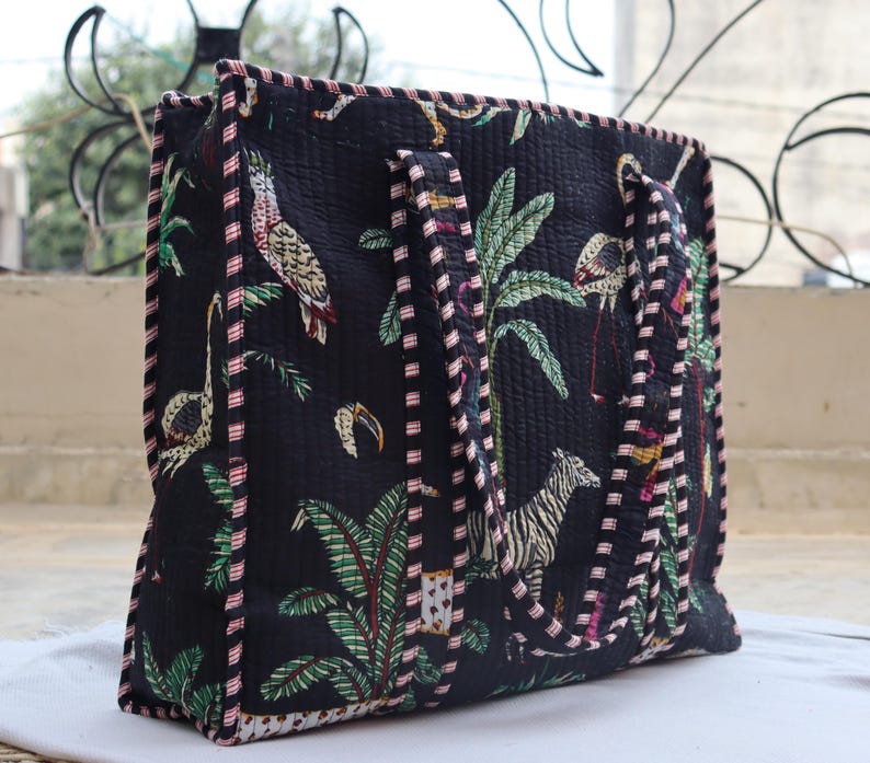 May include: Black tote bag with a quilted design featuring a jungle scene with parrots, flamingos, zebras, and palm trees. The bag has two handles and is trimmed with a pink and white striped border.