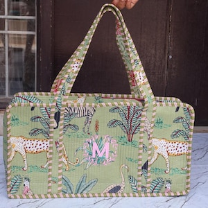 Puede incluir: Un bolso tote rectangular acolchado de tela verde claro con un patr&oacute;n repetitivo de leopardos, cebras y aves tropicales. El bolso tiene dos asas largas, un borde a rayas y una letra M bordada en color rosa en el panel frontal.