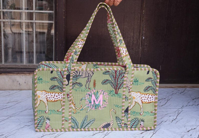 Puede incluir: Un bolso tote rectangular acolchado de tela verde claro con un patr&oacute;n repetitivo de leopardos, cebras y aves tropicales. El bolso tiene dos asas largas, un borde a rayas y una letra M bordada en color rosa en el panel frontal.
