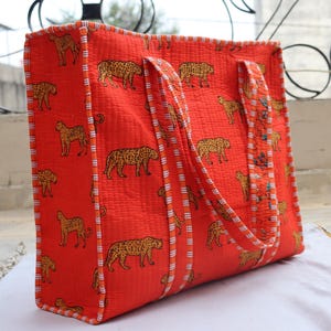 May include: A large, rectangular tote bag in a vibrant orange hue, adorned with a repeating pattern of tan leopards. The bag features sturdy handles and is trimmed with a striped border. This bag is perfect for carrying groceries or other items.