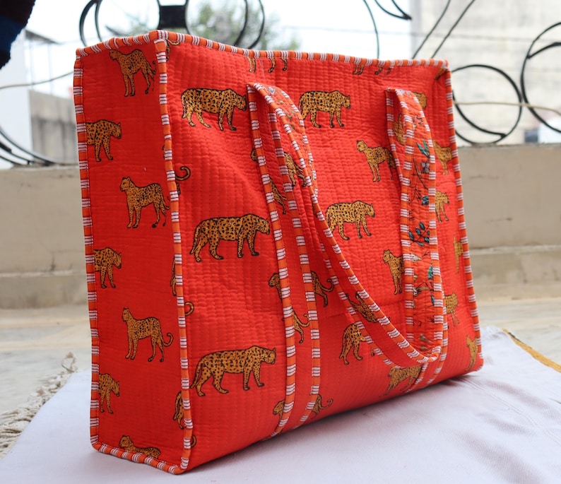 May include: A large, rectangular tote bag in a vibrant orange hue, adorned with a repeating pattern of tan leopards. The bag features sturdy handles and is trimmed with a striped border. This bag is perfect for carrying groceries or other items.
