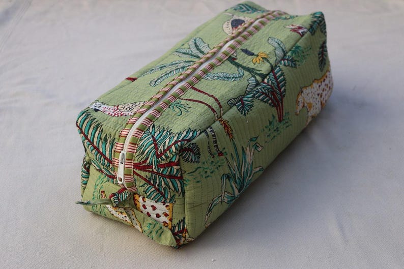 Puede incluir: Un neceser acolchado rectangular de tela verde claro con un patr&oacute;n repetitivo de plantas tropicales, hojas de palmera y animales moteados. El neceser incluye un cierre de cremallera blanco con un borde de tela a rayas rojas y blancas.