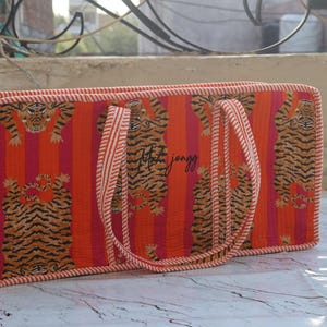 Puede incluir: Un bolso tote rectangular acolchado con un vibrante patr&oacute;n de rayas verticales naranjas y rosas con ilustraciones de tigres repetidas. El bolso incluye dos asas de tela a rayas a juego y un borde fino a rayas en todos los lados.