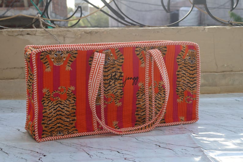 Puede incluir: Un bolso tote rectangular acolchado con un vibrante patr&oacute;n de rayas verticales naranjas y rosas con ilustraciones de tigres repetidas. El bolso incluye dos asas de tela a rayas a juego y un borde fino a rayas en todos los lados.
