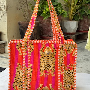 May include: A rectangular tote bag with a striking tiger print. The bag has a quilted design with orange, pink, and white stripes. The handles and edges are also striped. The tiger pattern is in shades of brown and black.