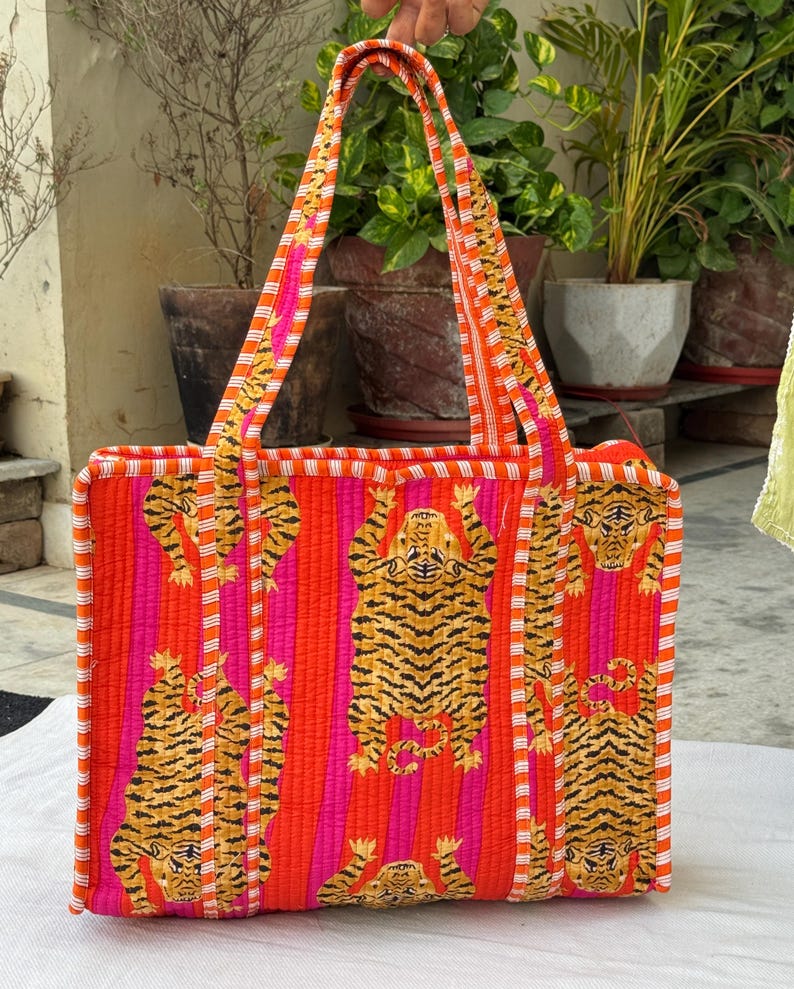 May include: A rectangular tote bag with a striking tiger print. The bag has a quilted design with orange, pink, and white stripes. The handles and edges are also striped. The tiger pattern is in shades of brown and black.