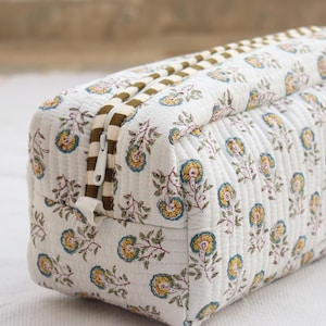 May include: A rectangular, quilted cosmetic bag with a floral pattern in yellow, blue, and brown on a white background. The bag has a brown zipper and a matching brown and white striped zipper pull. The bag is approximately 25 cm long.