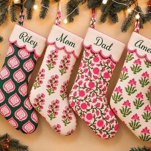 May include: Four fabric Christmas stockings with scalloped white cuffs featuring the names Riley, Mom, Dad, and Amelia embroidered in green. The stockings display various floral and leaf patterns in shades of pink, green, and cream fabric.