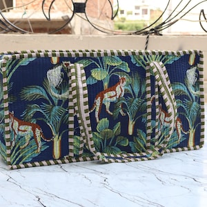 May include: A rectangular quilted tote bag featuring a dark blue background with a repeating pattern of orange and white cheetahs amidst green palm leaves. The bag has two matching fabric handles and green and white striped edge piping.