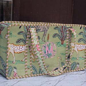 Puede incluir: Un bolso tote rectangular acolchado en verde claro con un estampado repetitivo de guepardos, cebras, flamencos y palmeras. El bolso tiene dos asas y un borde a rayas, con una letra M rosa bordada centrada dentro de una corona floral.
