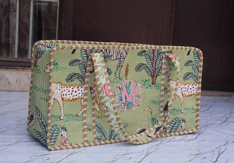 Puede incluir: Un bolso tote rectangular acolchado en verde claro con un estampado repetitivo de guepardos, cebras, flamencos y palmeras. El bolso tiene dos asas y un borde a rayas, con una letra M rosa bordada centrada dentro de una corona floral.